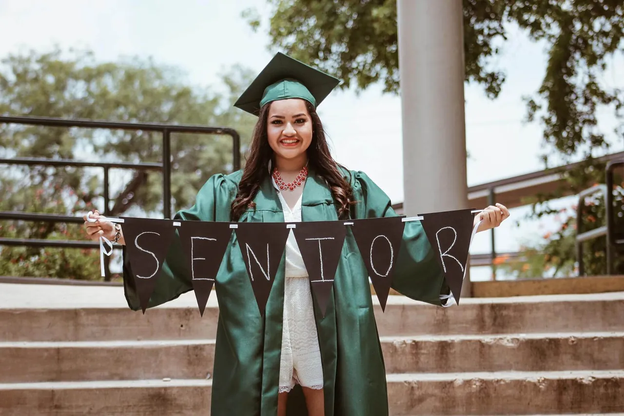 A graduating student in a green cap and gown holds a banner that spells "SENIOR" while standing on outdoor steps, contemplating the future as she thinks about seeking counseling in Sugar Land, TX.
