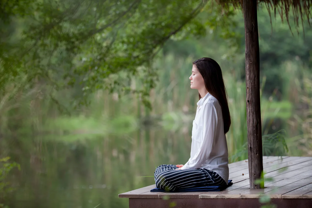 A woman sits cross-legged on a wooden deck by a serene lake, meditating. She is wearing a white shirt and striped pants, surrounded by lush green foliage, finding inner peace just like you'd hope to with an anxiety therapist near you.