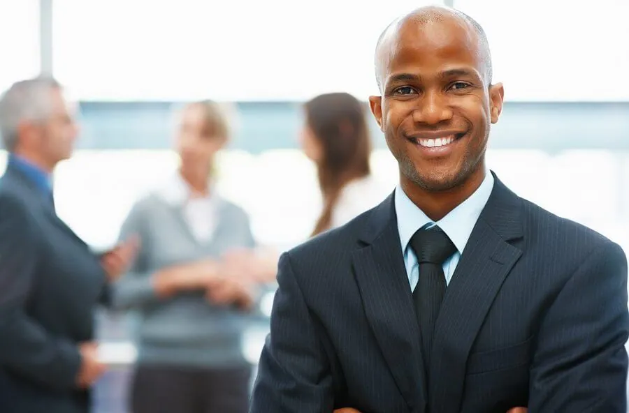 A person in a suit smiles confidently with their arms crossed, embodying professionalism and ease. In the background, three blurred individuals engage in discussion, perhaps seeking to find a therapist near Sugar Land for guidance and support.
