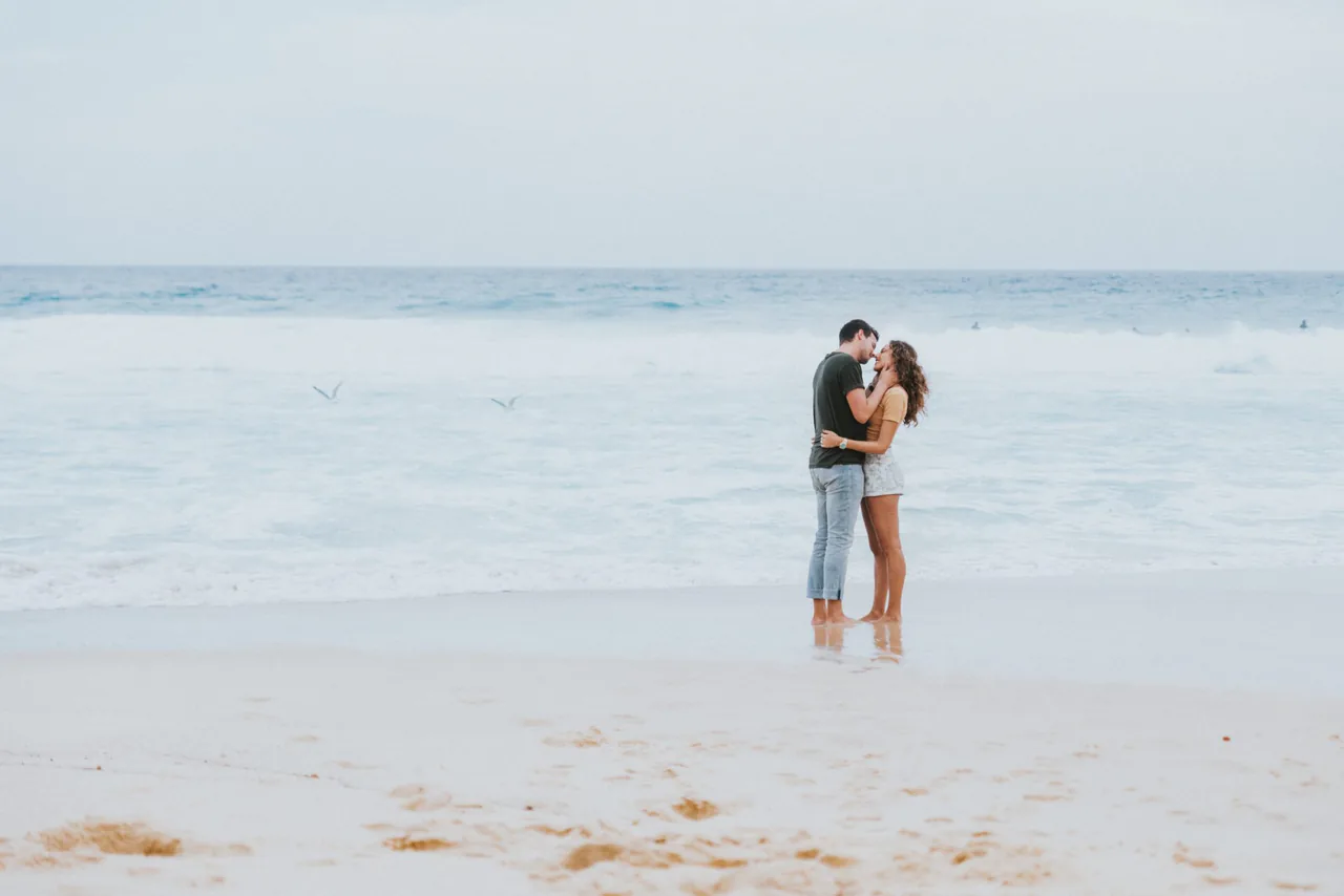 A couple embracing on a sandy beach with gentle waves in the background under an overcast sky, perhaps finding solace similar to counseling in Sugar Land, TX.
