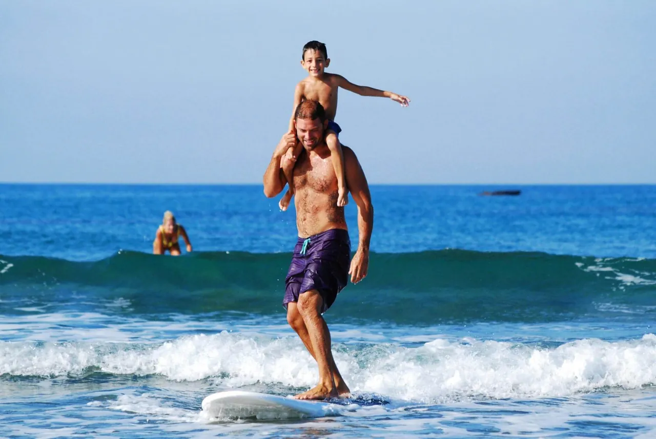 A man surfing the ocean waves with a boy on his shoulders, both in swimwear and smiling, captures a joyful escape. In the background, another surfer glides smoothly. For those seeking balance like this in life, counseling in Sugar Land, TX offers support and guidance along the way.