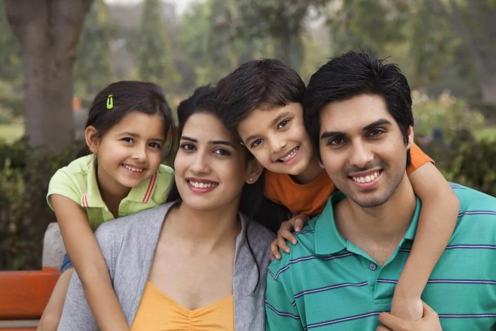 A family of four sits on a park bench. The mother and father are seated with their two children leaning against them, all smiling, perhaps after visiting the best therapists near them. The backdrop features trees and lush greenery, providing a tranquil setting for togetherness.