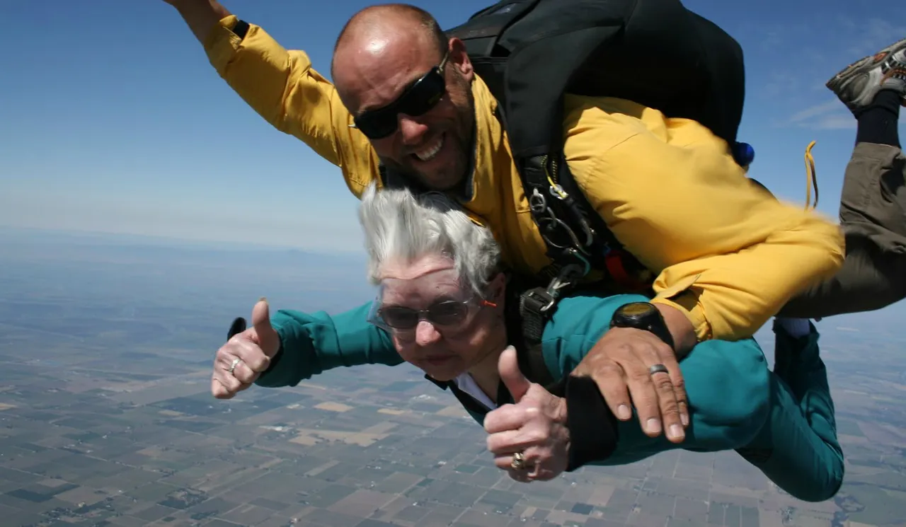 A tandem skydiving pair, an older woman in a blue jumpsuit and her instructor in yellow, gives thumbs up while descending over a sunny landscape. It's as exhilarating as finding the right counselor in Sugar Land, TX, to guide you through life's challenges with confidence and ease.