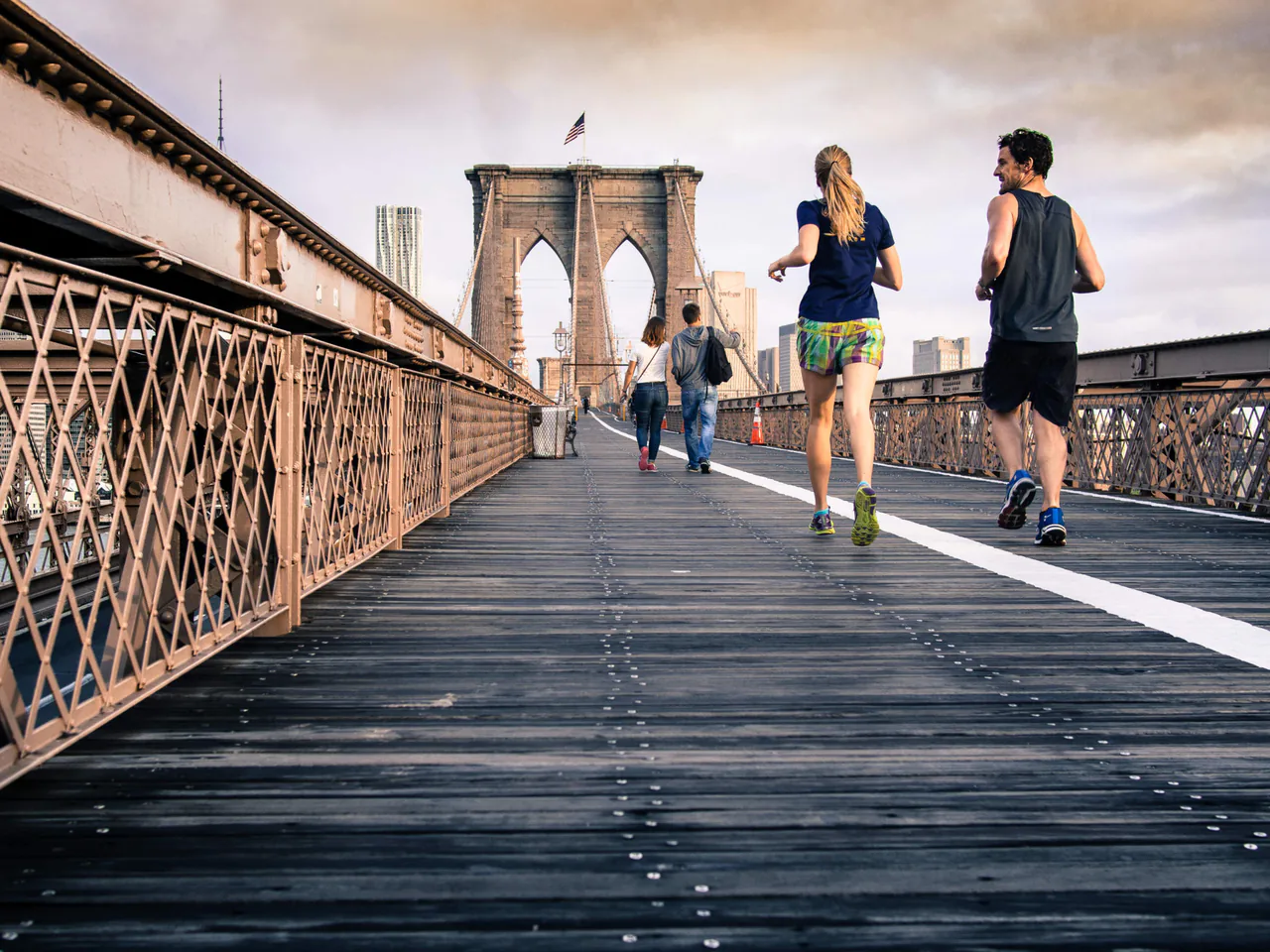 Two people jog on the wooden path of the Brooklyn Bridge, with others walking ahead under a cloudy sky, as if contemplating where to find a therapist near Sugar Land for some much-needed counseling.